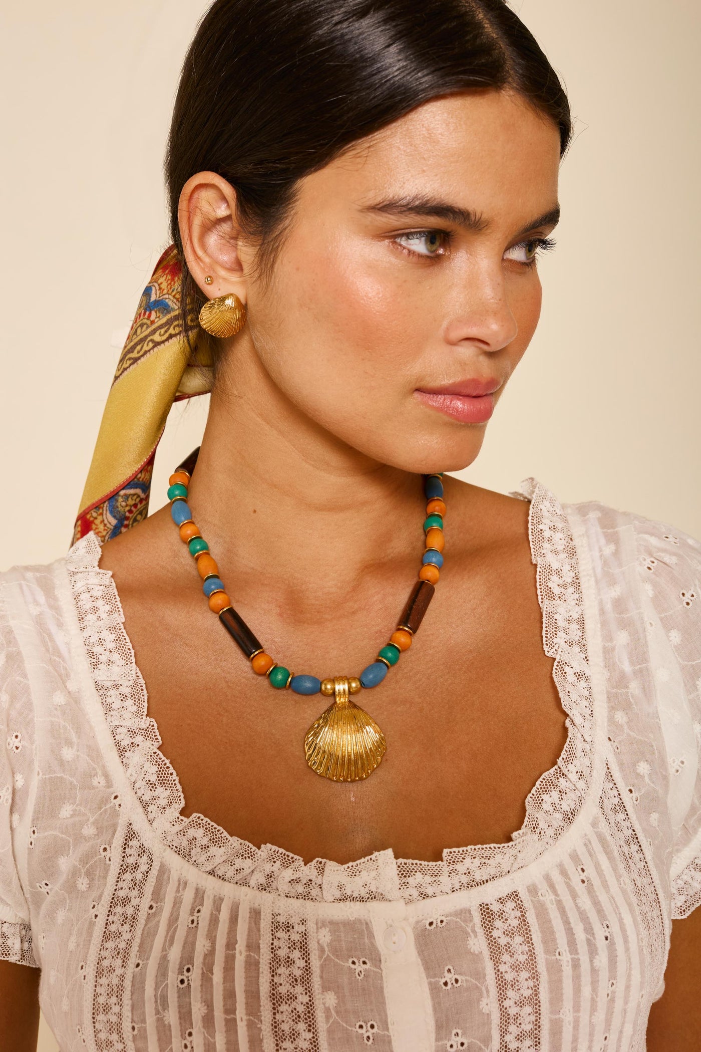 Woman with dark hair tied back in a colorful scarf, wearing a white lace top, gold earrings, and the Darielle - Multi necklace featuring turquoise, orange, and wooden beads with a gold-plated shell pendant, looking to the side.