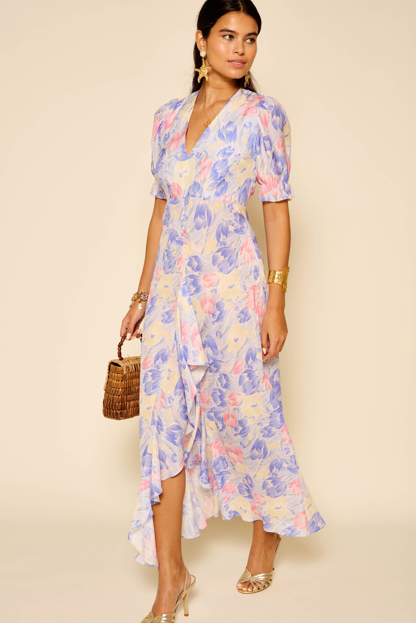 A woman in the Ariel - Tulip Fields Blue dress, a vintage-style pastel floral midi with short sleeves and V-neckline, accessorized with gold jewelry, strappy heels, and a woven bag, poses against a beige background—ideal for special occasions.
