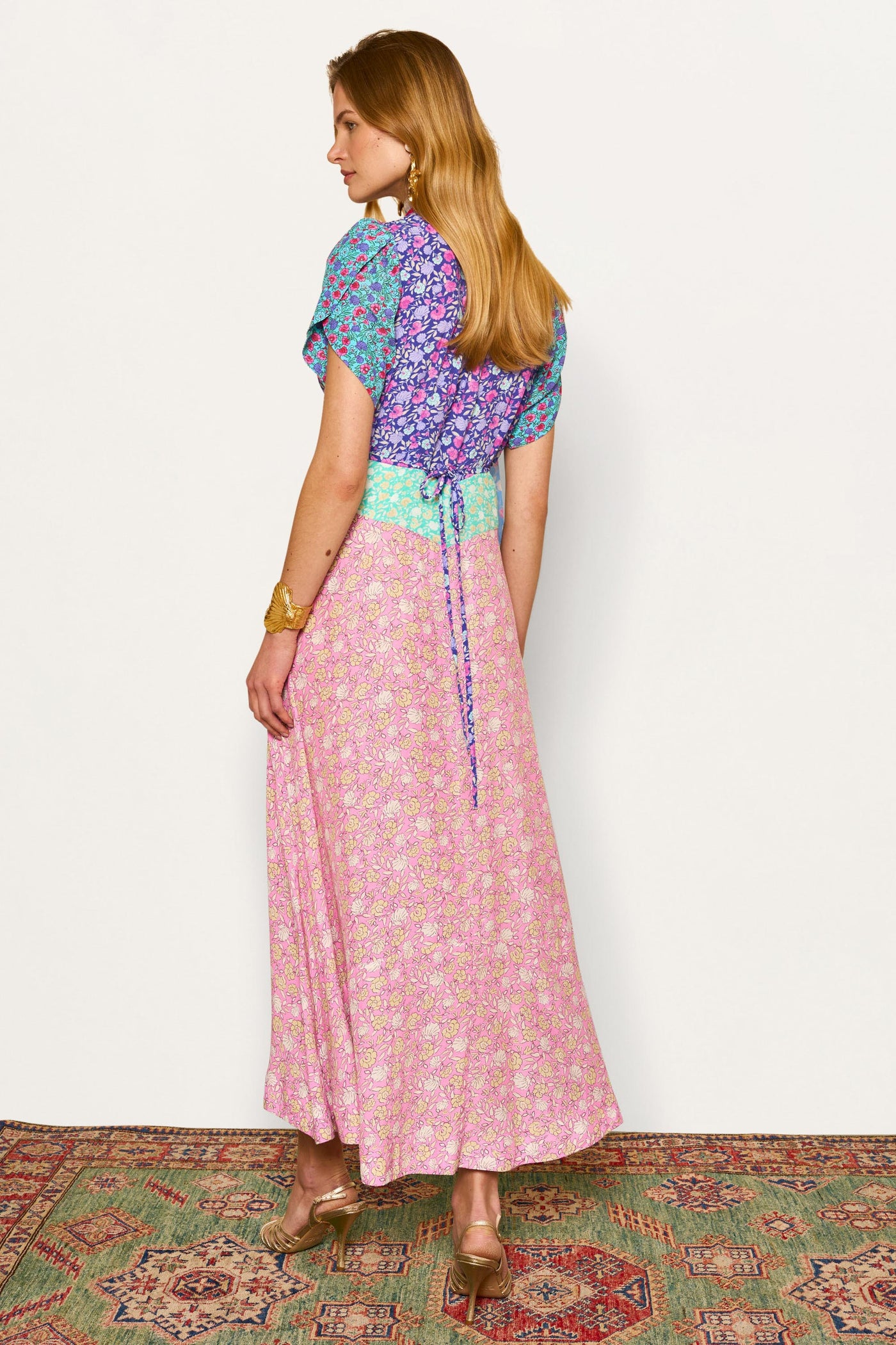 A woman with long light brown hair and gold jewelry stands on a patterned rug, facing away, wearing the Novara - Shell Pansy Mix Multi dress with a blue-purple top, green waistband, pink skirt, curved sleeves, and gold heels.