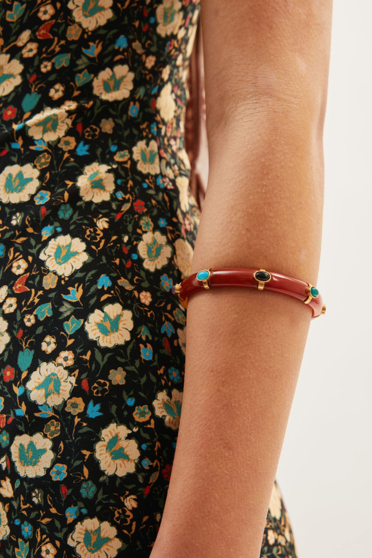 A person in a black floral dress with cream, teal, and yellow designs wears the Sabella - Tortoise, a gold-plated arm cuff with turquoise and black stones, on their upper arm.