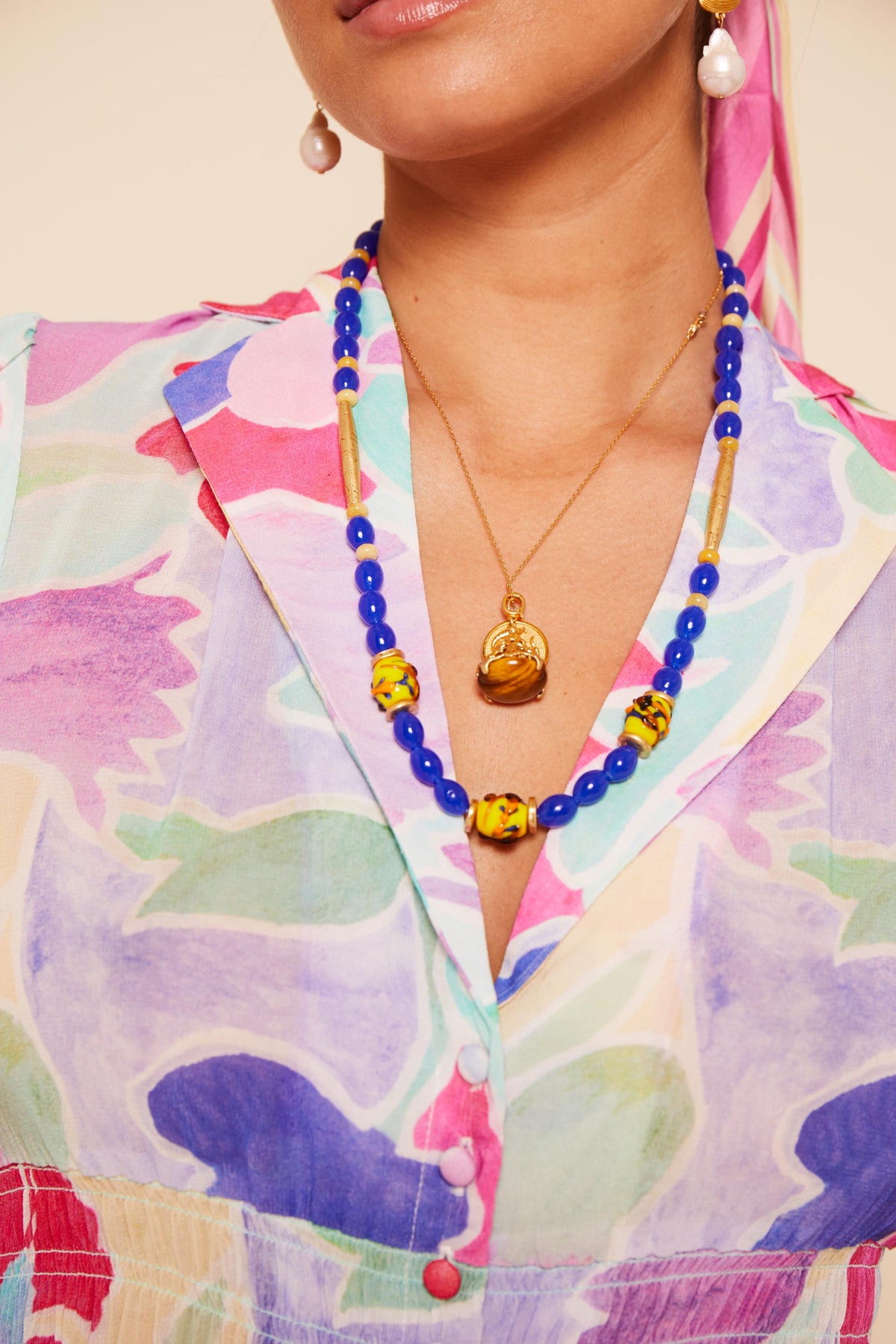 A woman wears a colorful floral blouse, the Denali - Multi blue and yellow beaded necklace, a gold pendant necklace, pearl drop earrings, and a pink headscarf. The focus is on her accessories and upper torso.