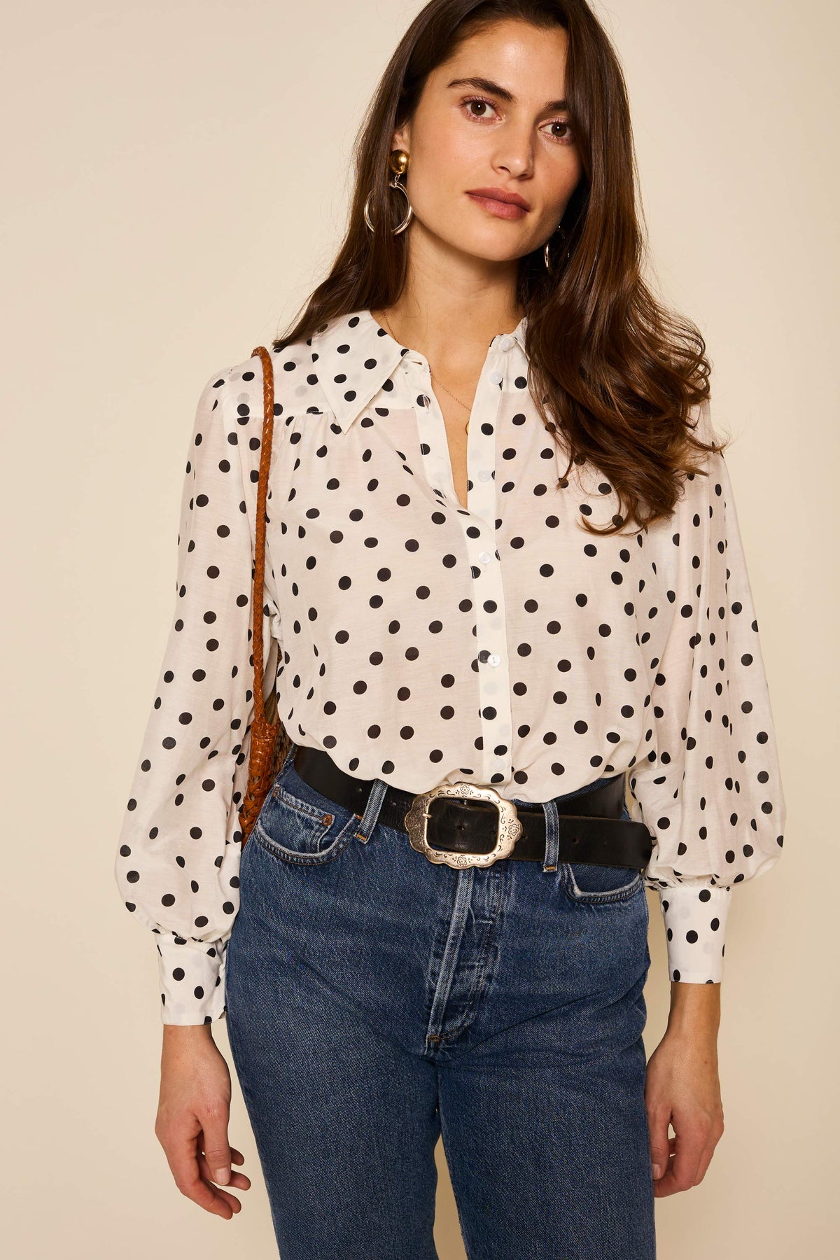 Woman with long brown hair wears the Blake - Bohemia Spot Top, featuring an exaggerated collar and black polka dots, paired with blue jeans, a wide black belt with a decorative buckle, and gold hoop earrings against a plain background.