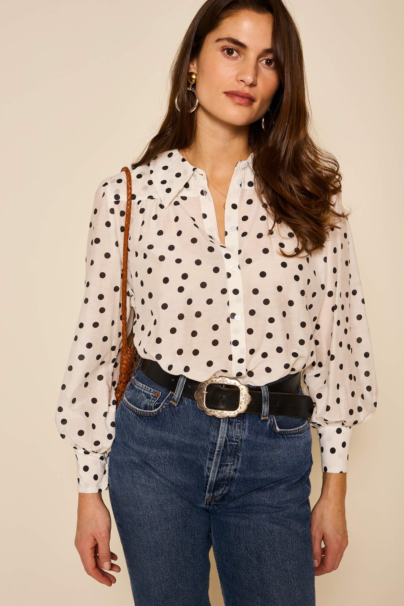 Woman with long brown hair wears the Blake - Bohemia Spot Top, featuring an exaggerated collar and black polka dots, paired with blue jeans, a wide black belt with a decorative buckle, and gold hoop earrings against a plain background.