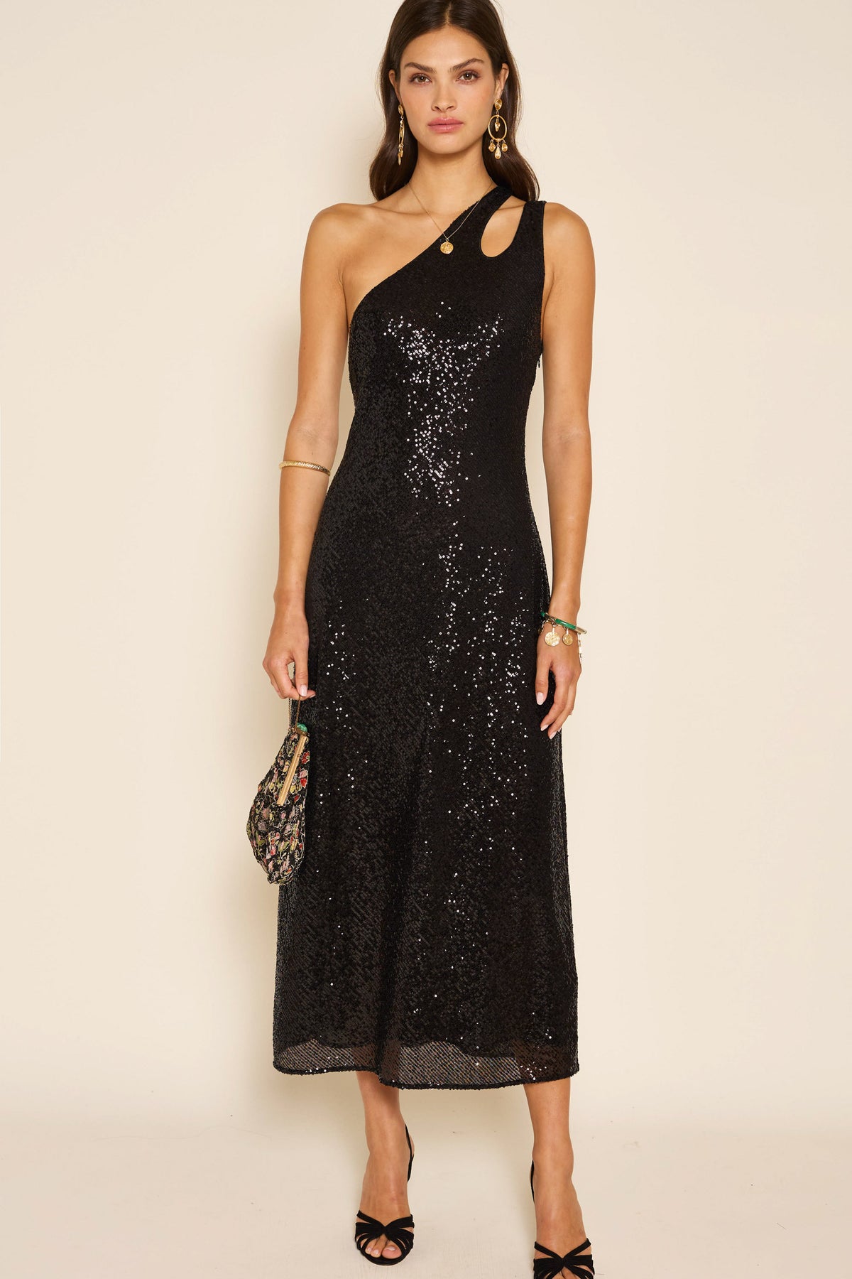A woman showcases the Avena - Sequin Black, a black sequin maxi dress featuring a one-shoulder, asymmetric silhouette with chic cutout detailing. She completes her elegant party look with black heels and a small patterned clutch, standing poised against a plain backdrop.