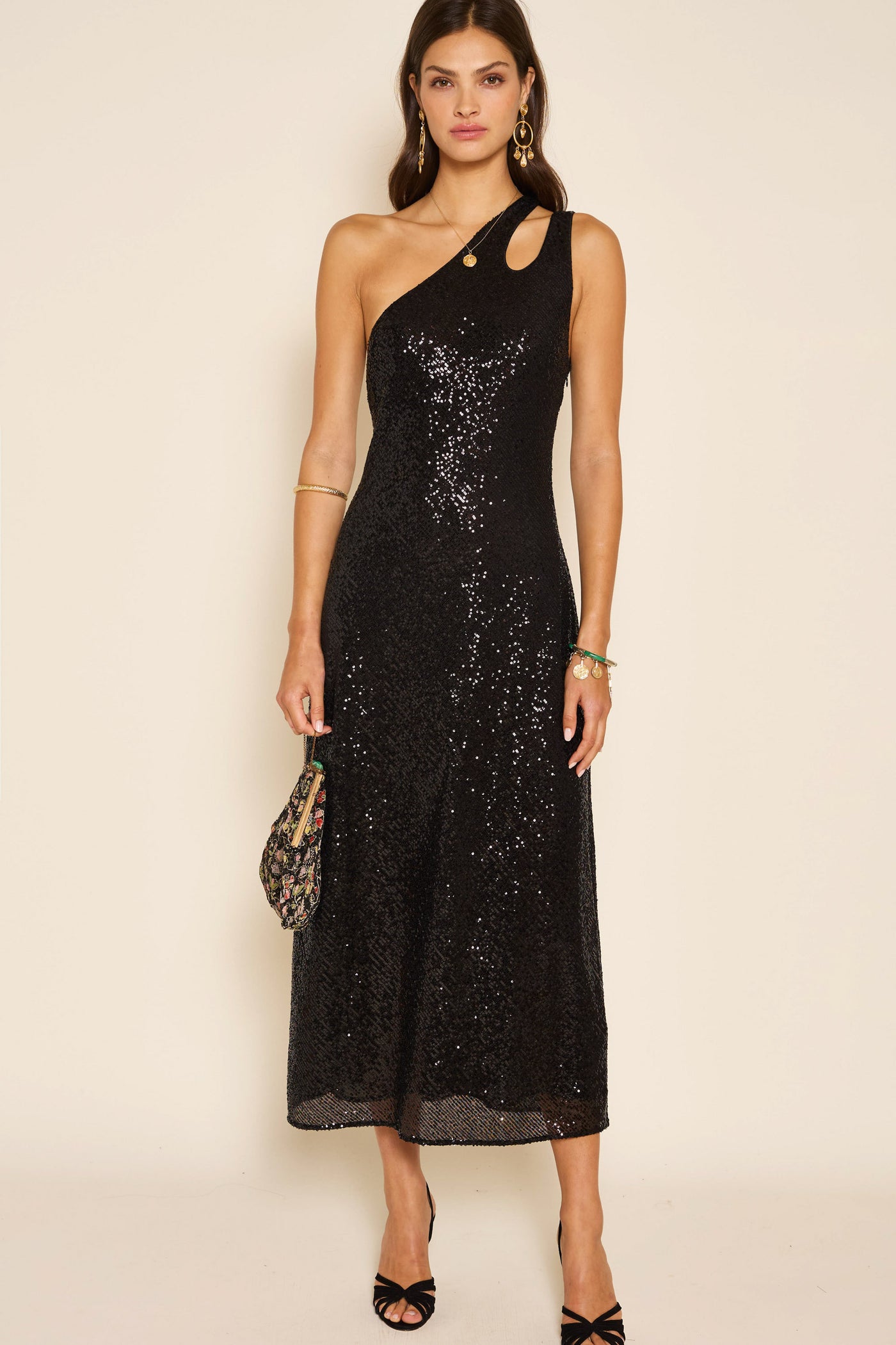 A woman showcases the Avena - Sequin Black, a black sequin maxi dress featuring a one-shoulder, asymmetric silhouette with chic cutout detailing. She completes her elegant party look with black heels and a small patterned clutch, standing poised against a plain backdrop.