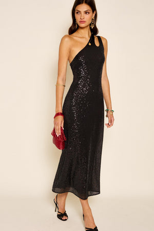 A woman wears the Avena - Sequin Black one-shoulder party dress, paired with black heels, gold earrings, a gold bangle, and a red textured clutch. She stands against a plain light background.