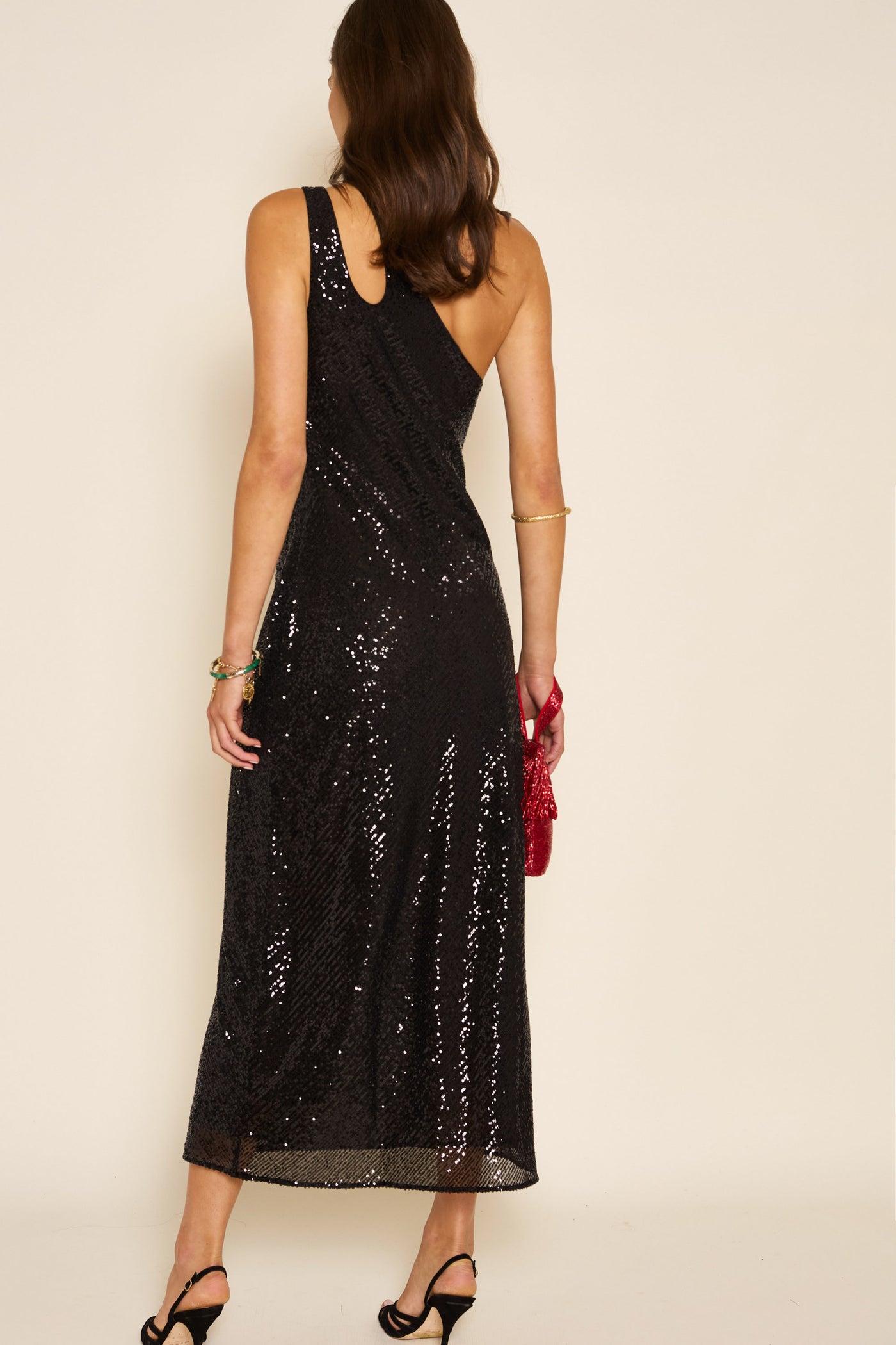 A woman stands facing away, wearing the chic Avena - Sequin Black maxi dress, featuring an asymmetric cut and adorned with black sequins. She pairs it with elegant black heels and carries a red clutch in her right hand. Her loose hair and a gold bracelet complete the look, while the plain background perfectly showcases this stunning party dress.