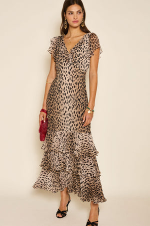 A woman with long brown hair and a neutral expression stands against a plain background wearing the Amelle - Antelope Spot Black, a 1930s-inspired, v-neck, ankle-length leopard print dress with fluted sleeves.