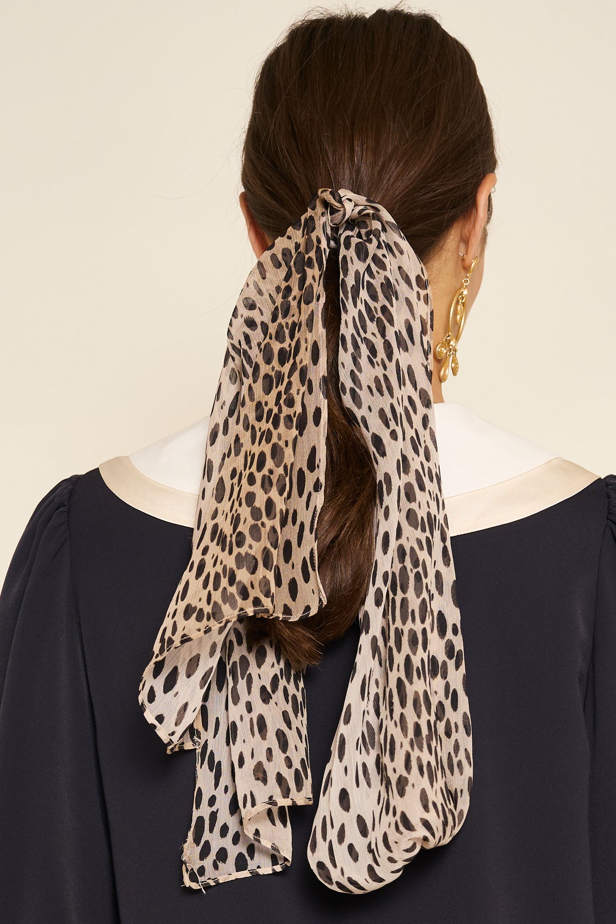 A woman with brown hair in a low ponytail and gold drop earrings wears a dark top with cream trim. Seen from behind, she accessorizes with the Julien Scarf Antelope Spot Black—a long, sheer, crinkled-georgette antelope-print scarf.