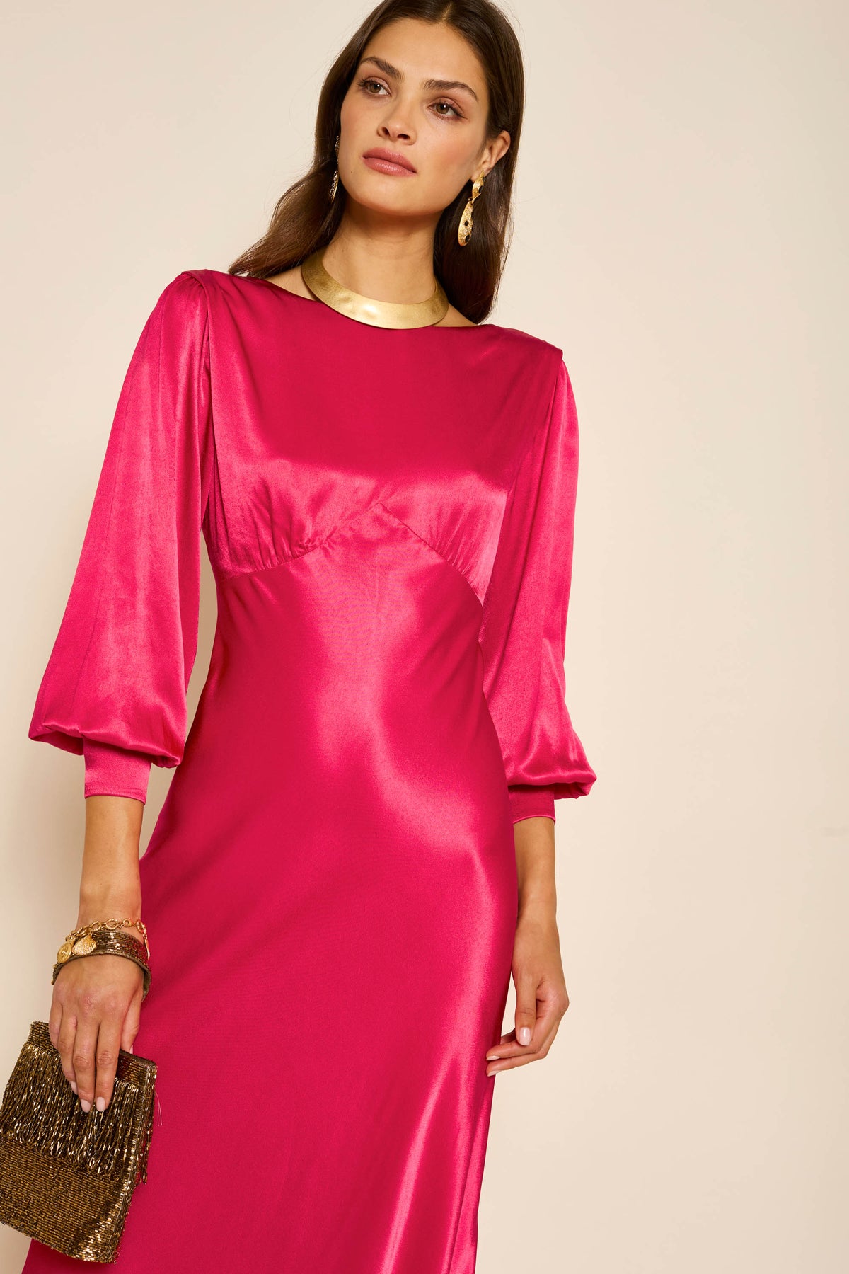 A woman in the "marni - raspberry" vintage-inspired satin dress holds a brown beaded clutch. She has long brown hair, wears gold earrings and a necklace, standing against a plain light background.
