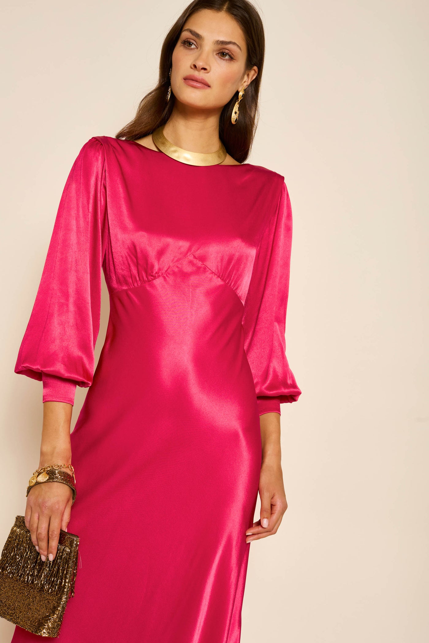 A woman in the "marni - raspberry" vintage-inspired satin dress holds a brown beaded clutch. She has long brown hair, wears gold earrings and a necklace, standing against a plain light background.