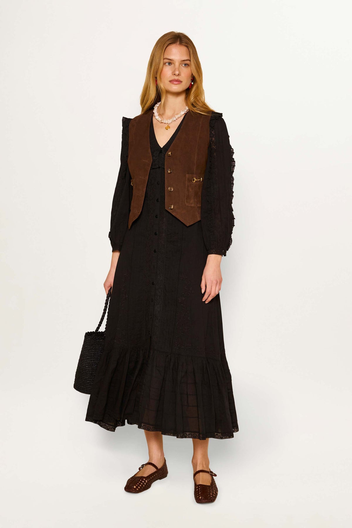 A woman wears a long black dress with a ruffled hem, a vintage waistcoat, and Rikki - Tan shoes. She carries a black woven bag, accessorizes with a pearl necklace, and her long hair is loose and center-parted.