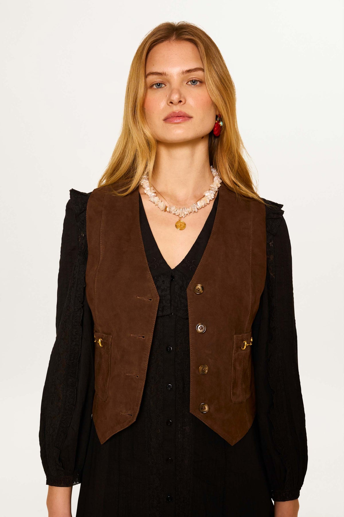 A woman with long blonde hair wears the Rikki - Tan vintage brown suede waistcoat over a black lace dress, accessorized with a chunky beaded necklace and red earrings, standing against a plain white background.