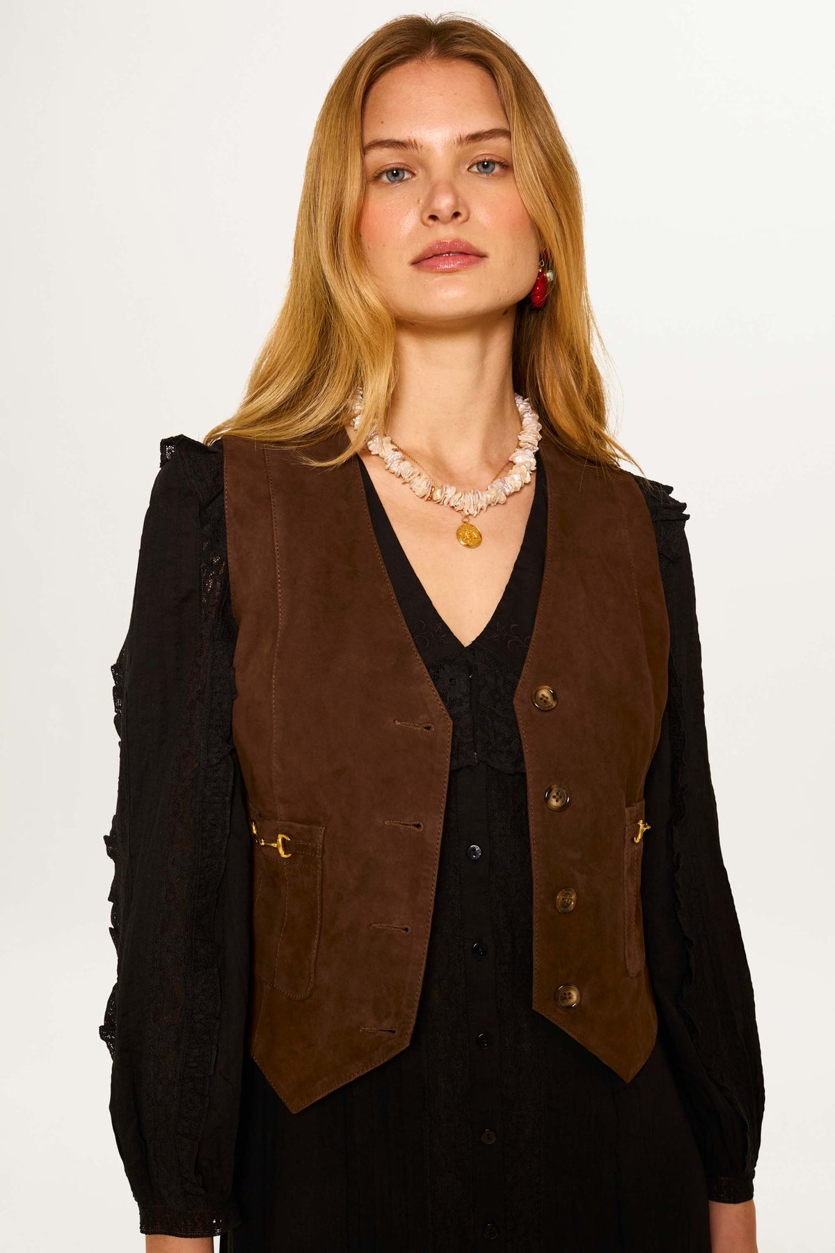 A woman with long blonde hair wears the Rikki - Tan suede waistcoat over a black dress, accessorized with a shell necklace and gold pendant, standing against a plain light background.