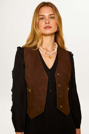 A woman with long blonde hair wears the Rikki - Tan suede waistcoat over a black dress, accessorized with a shell necklace and gold pendant, standing against a plain light background.