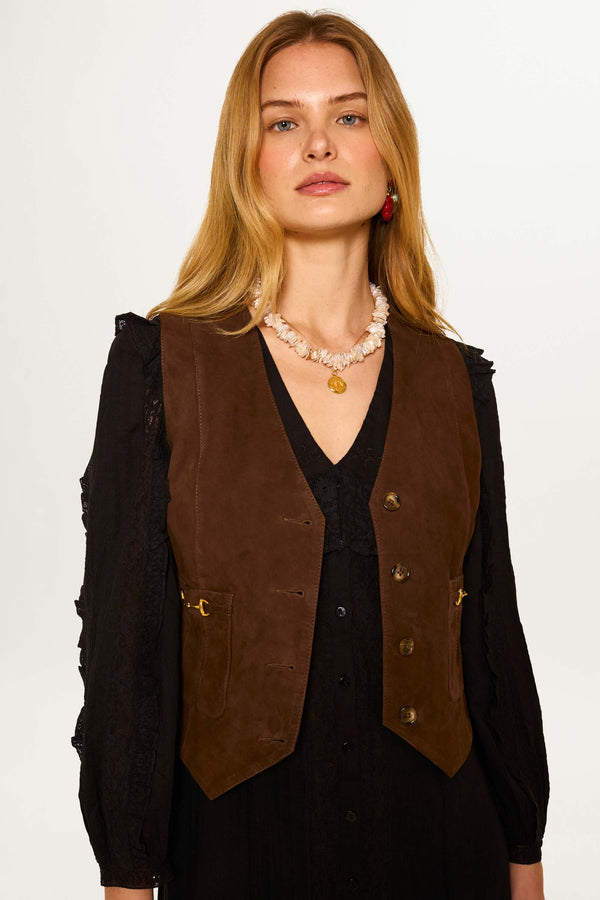 A woman with long blonde hair wears the Rikki - Tan suede waistcoat over a black dress, accessorized with a shell necklace and gold pendant, standing against a plain light background.