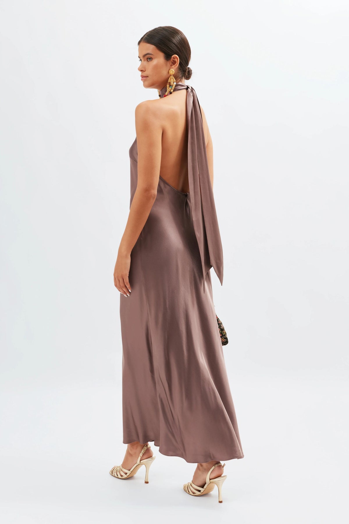 A woman in a long, open-back Maura - Mink gown with a scarf-like halter neck tie and cream high-heeled sandals stands sideways against a plain white background.