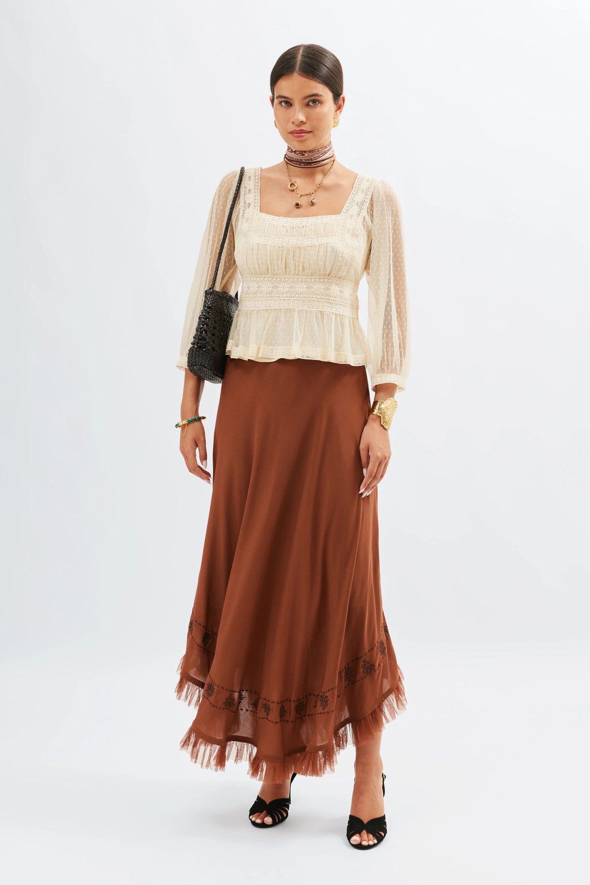 A woman poses against a white background wearing the Tyla - Vintage Cream blouse with sheer Swiss dot tulle sleeves, styled with layered necklaces, bracelets, a choker, black purse, black sandals, and an ankle-length brown skirt.