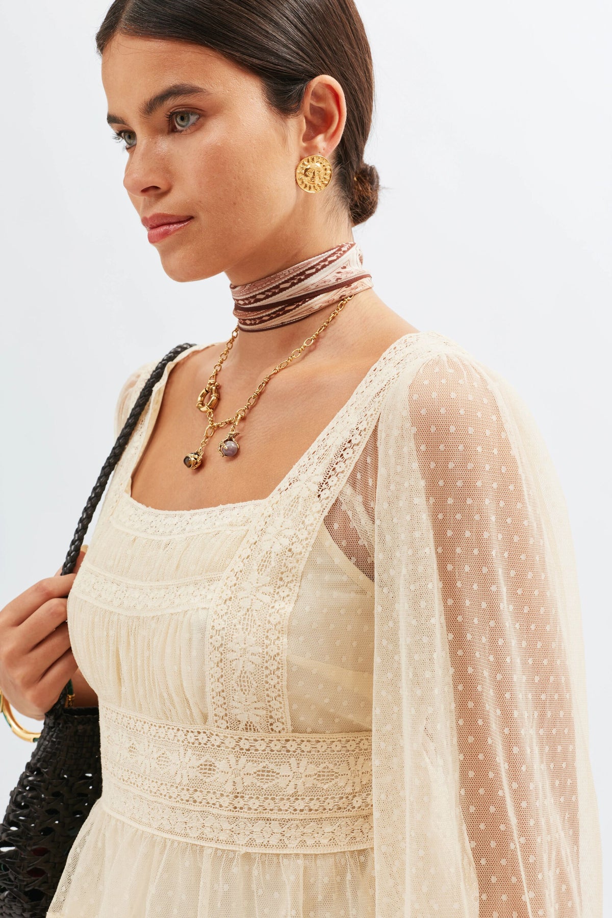 Wearing the Tyla - Vintage Cream dress with sheer Swiss dot tulle sleeves, gold earrings, layered necklaces, and a brown ribbon choker, a woman stands in profile holding a black bag over her shoulder.