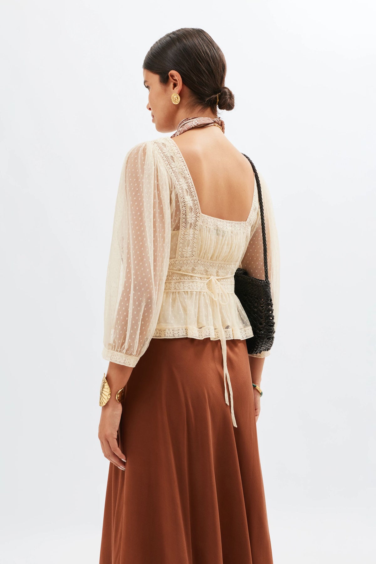 A woman with dark hair in a low bun wears the Tyla - Vintage Cream square neck lace blouse with sheer sleeves, styled with a brown skirt, black woven shoulder bag, and gold jewelry against a plain light background.