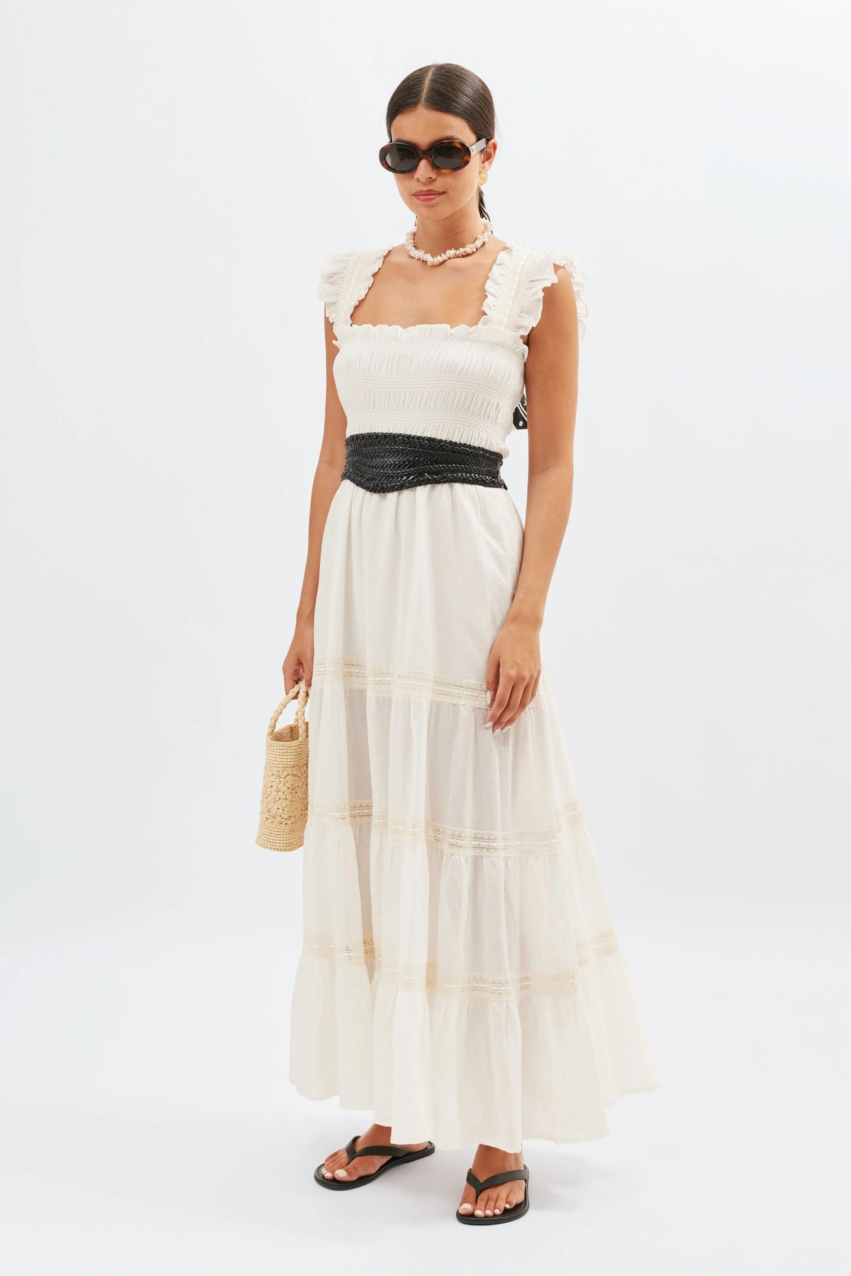 A woman wearing the Aubrie - Ivory white, ruffled nightdress styled as a maxi dress, cinched with a black belt and paired with black sandals and sunglasses, holds a small woven bag while standing against a plain white background.