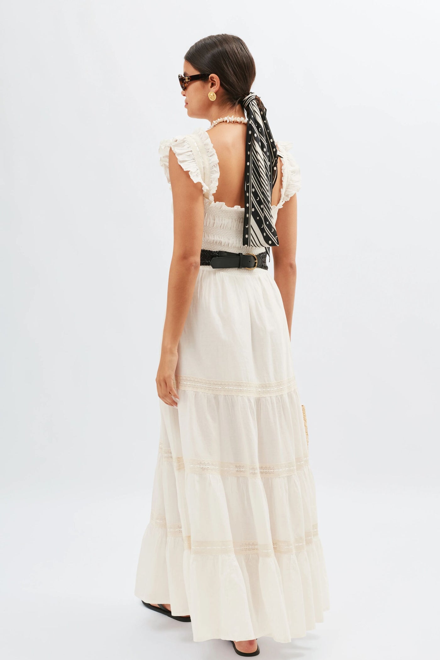 A woman wearing the Aubrie - Ivory, a long, white, 70s-inspired nightdress with ruffled straps, stands facing away. She has a black and white scarf tied in her hair, sunglasses, gold earrings, a black belt, and black sandals.