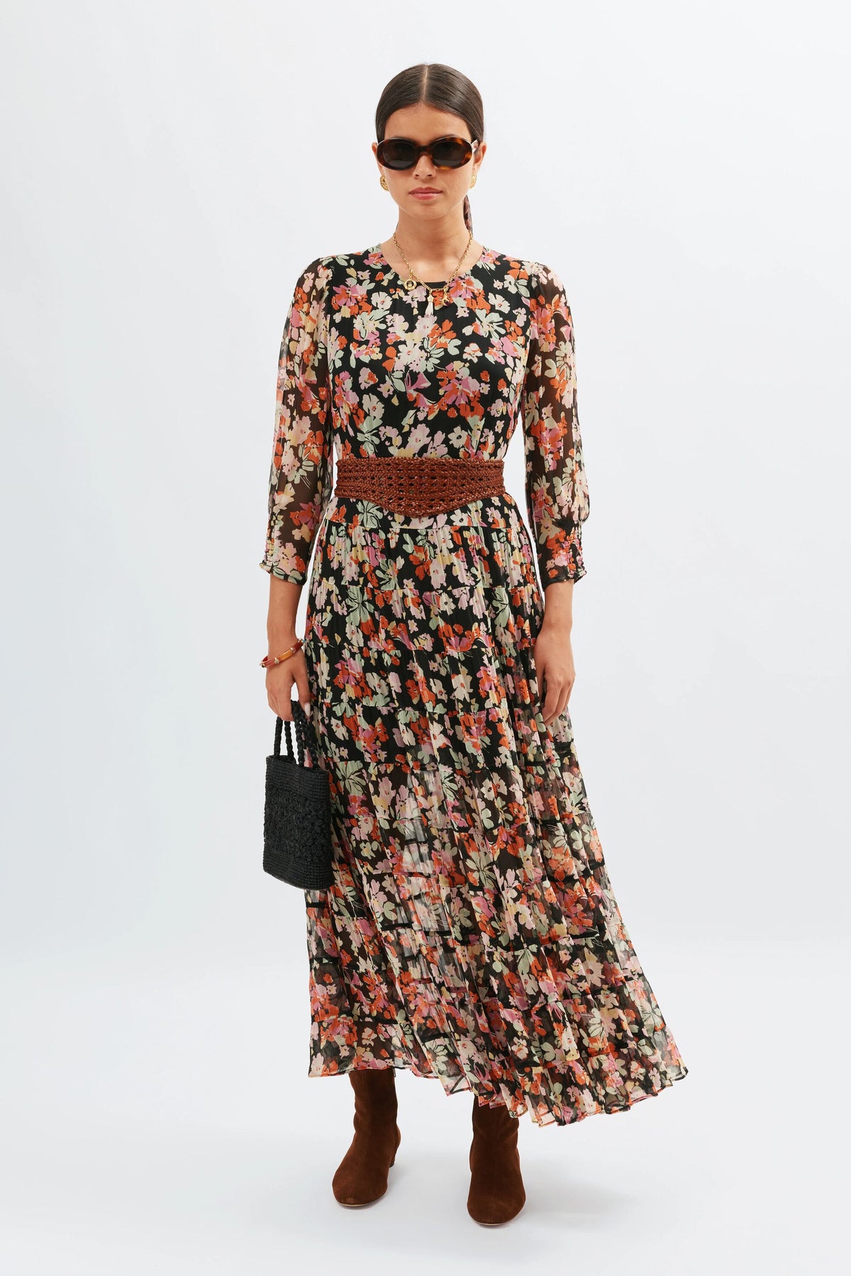 A woman wearing the Kristen - Primrose Floral Deep Navy dress, styled with brown ankle boots, a woven belt, sunglasses, and a small black handbag stands against a plain white background.