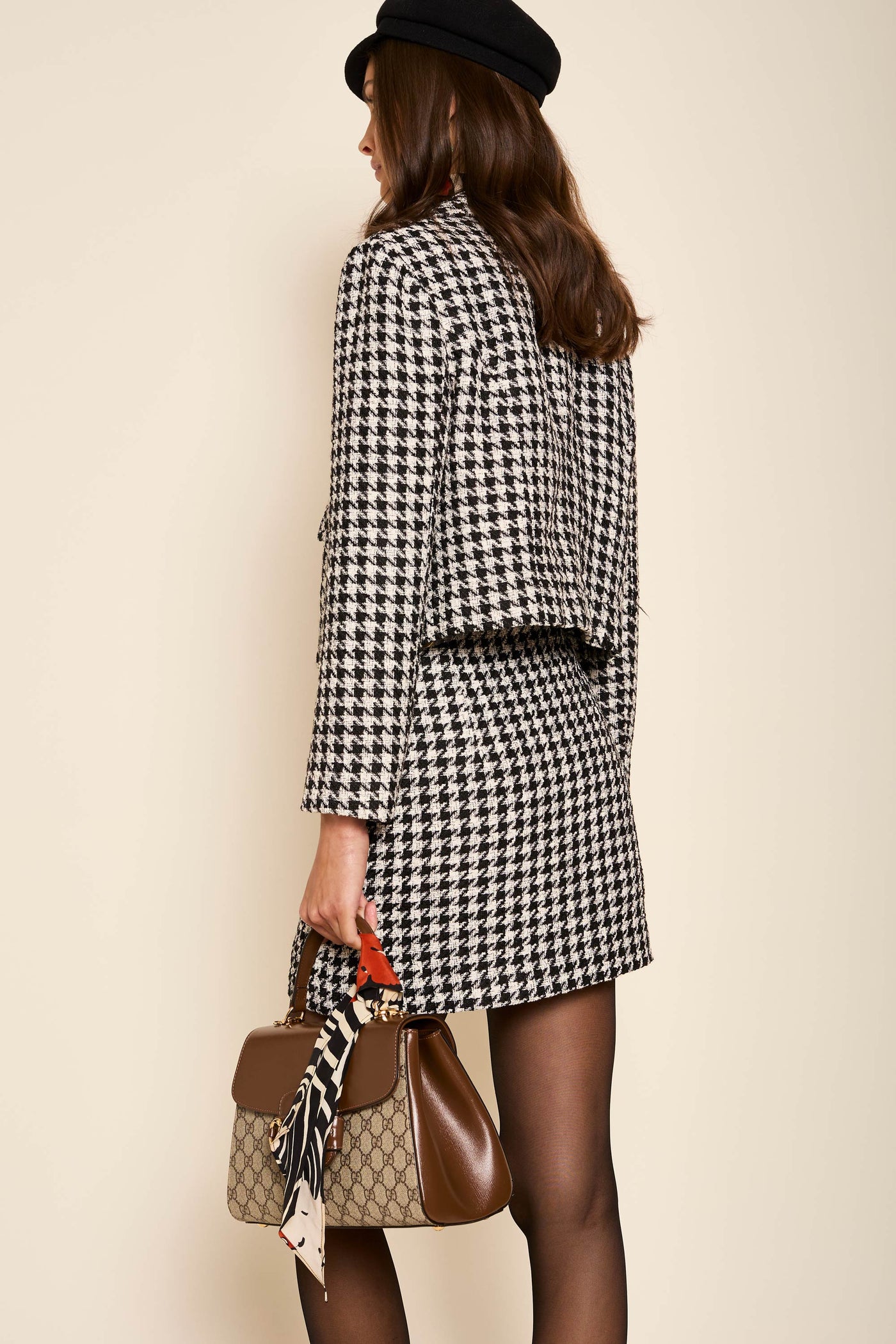 A woman faces away in true noughties style, pairing the Mariah - Houndstooth Mono mini skirt with a black beret, long blazer, black tights, and a brown handbag accented by a printed scarf.
