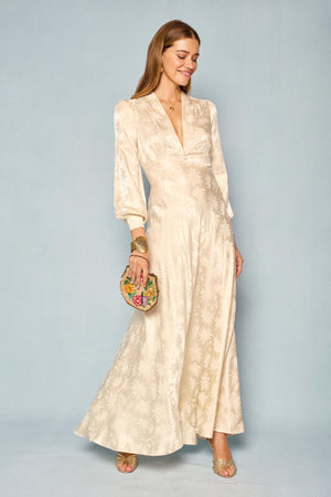 A woman stands smiling against a plain light blue background, wearing the Emory - Daisy Jacquard Champagne dress, a cream long-sleeve satin gown with a delicate floral pattern reminiscent of vintage wedding attire. She accessorizes with gold sandals, elegant jewelry, and holds a round clutch embroidered with flowers.