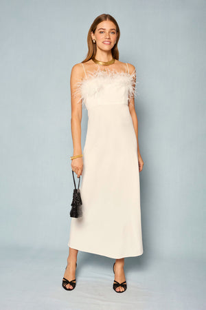 A woman wears the Selene - Ivory sleeveless white dress featuring feather trim at the top, paired with black heels, gold jewelry, and a small black clutch, standing against a plain light blue background.