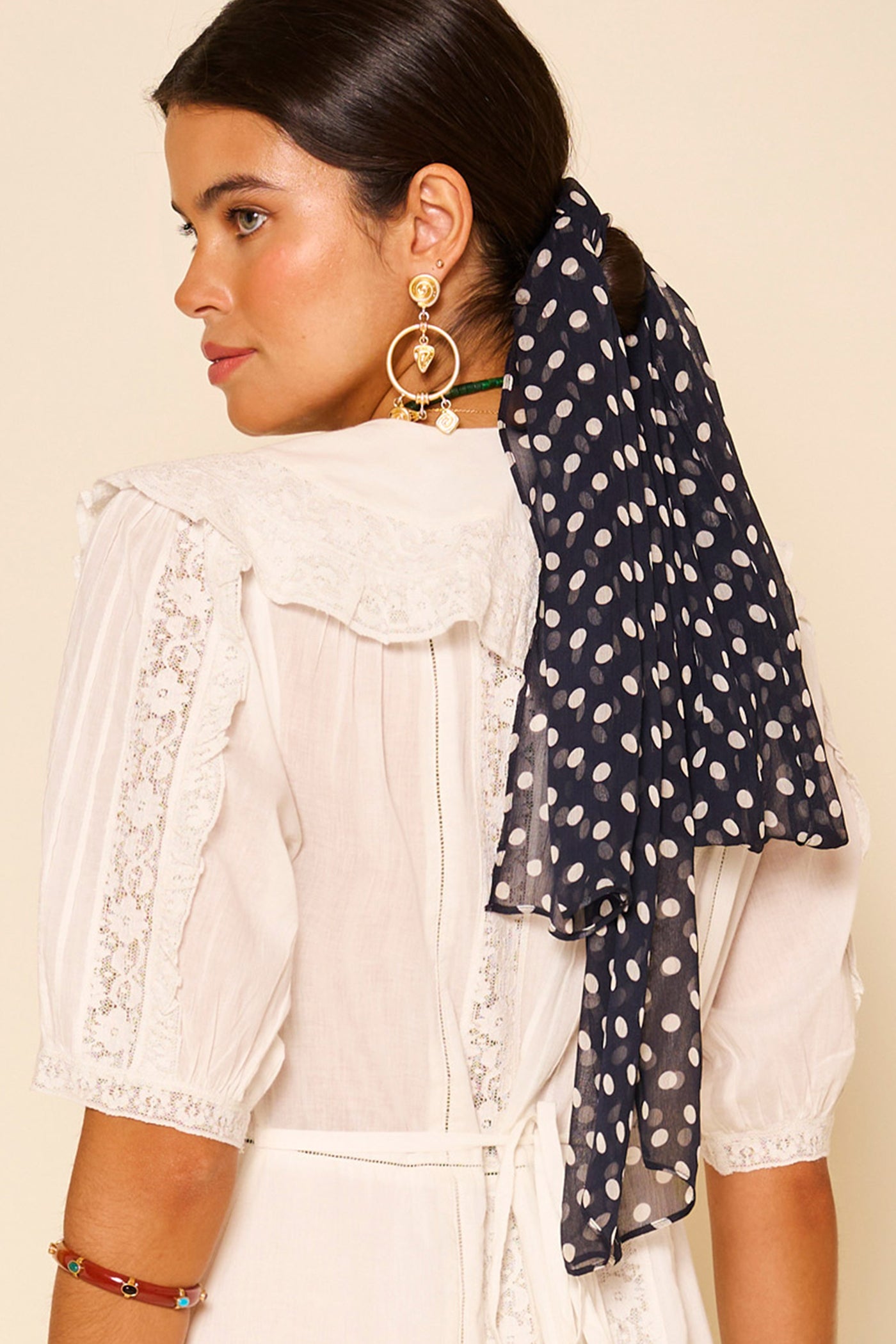 A woman with dark hair in a low ponytail, tied with the Julien - Emma Spot Deep Navy scarf, wears a white lace-trimmed dress and gold statement earrings, looking over her shoulder.