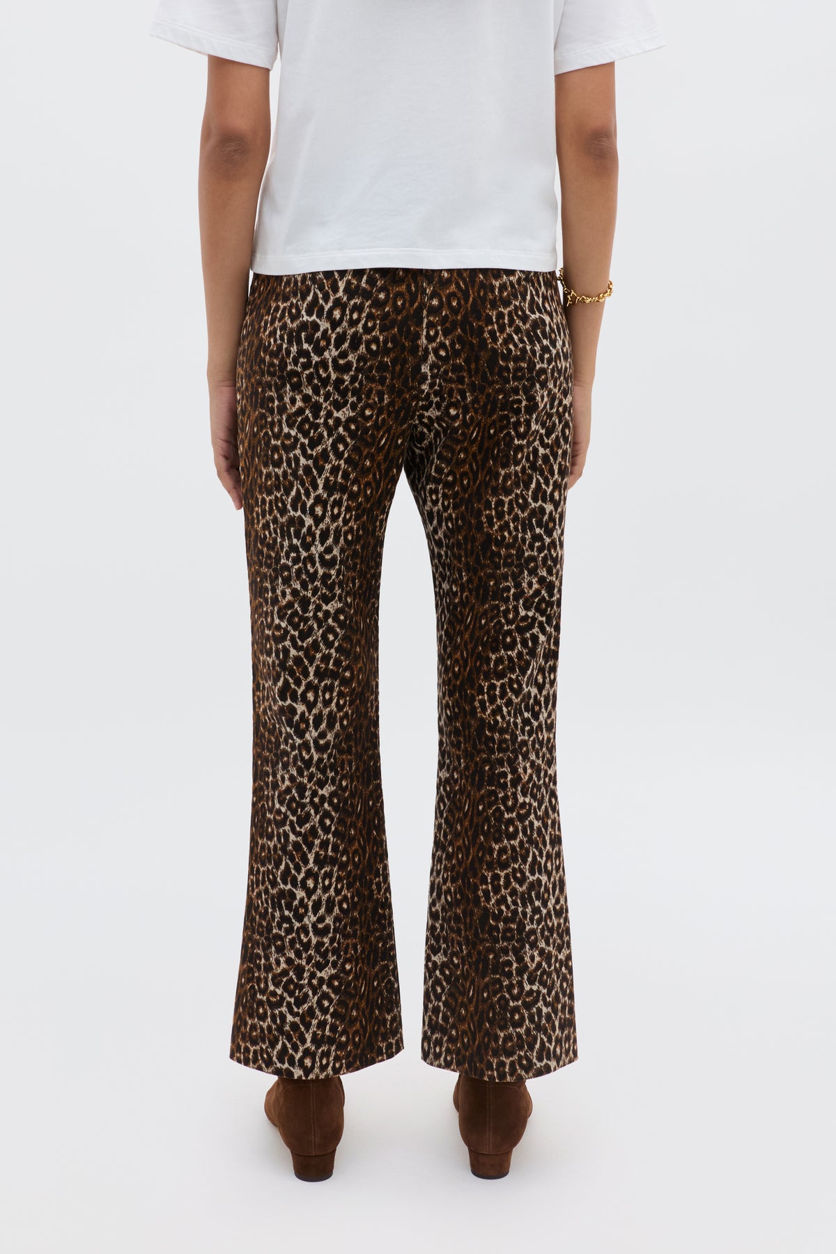 A person seen from behind wears Olwen - Lyra Leopard Black trousers, a white short-sleeve top, brown shoes, and a gold bracelet against a plain white background.