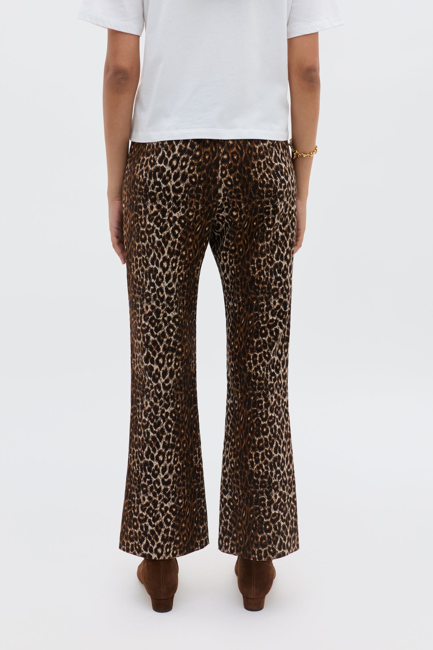 A person seen from behind wears Olwen - Lyra Leopard Black trousers, a white short-sleeve top, brown shoes, and a gold bracelet against a plain white background.