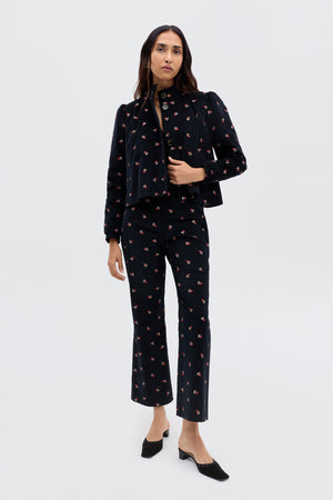 A woman stands against a plain background wearing the Olwen - Dandy Embroidery Deep Navy set, featuring vintage-inspired tailored trousers and a matching top with black floral embroidery, paired with black low-heeled shoes.