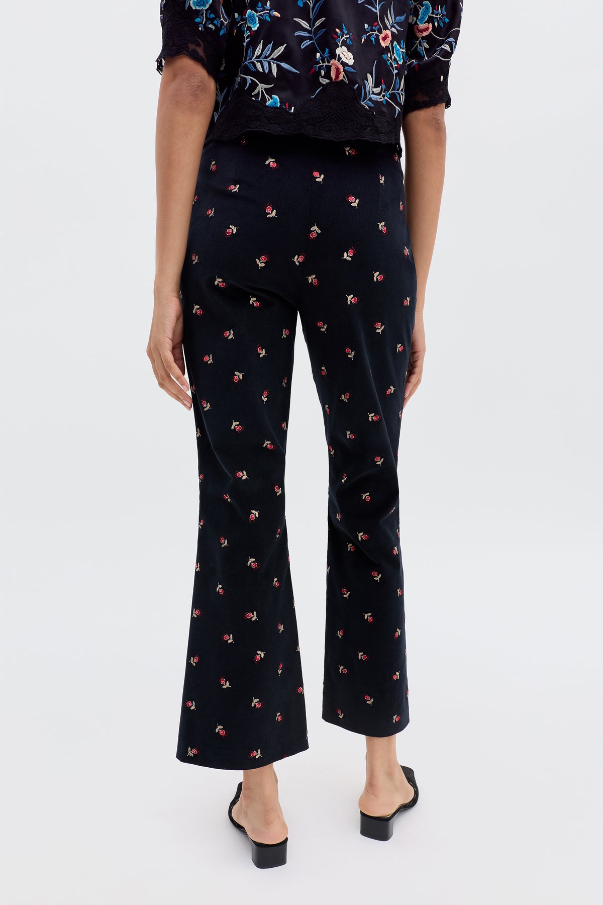 Wearing the Olwen - Dandy Embroidery Deep Navy top with blue and red floral embroidery, floral print trousers featuring a small red pattern, and black open-back heeled shoes, against a plain light background.
