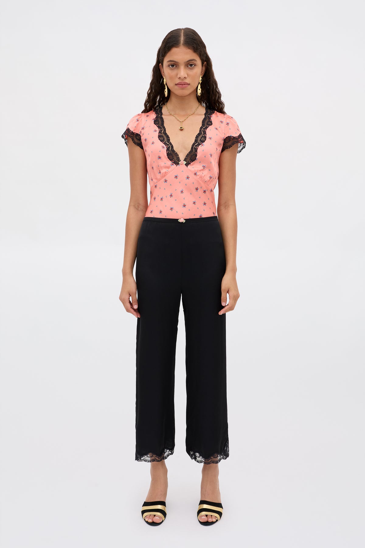 A woman with long wavy hair stands against a plain background, wearing the Mireille - Black wide-leg pants with lace at the hems, paired with a pink top featuring black lace trim and black sandals.