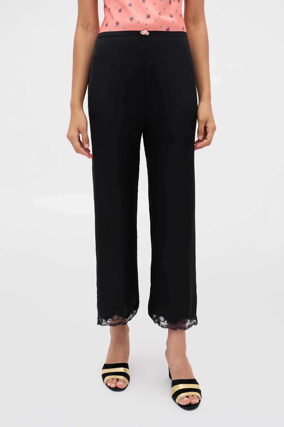 Wearing Mireille - Black straight-leg pants with lace-trimmed hems, a peach sleeveless top adorned with small star patterns, and open-toe heels featuring black and beige straps. Upper body and head are out of frame.