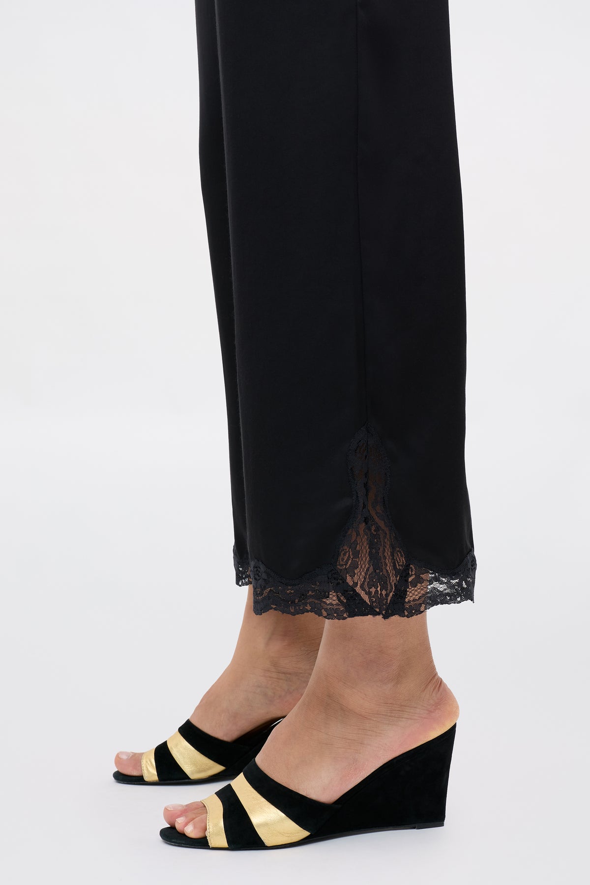 A person wearing the Mireille - Black satin pants with lace-trimmed hems and black wedge sandals featuring gold stripes, standing on a light background.