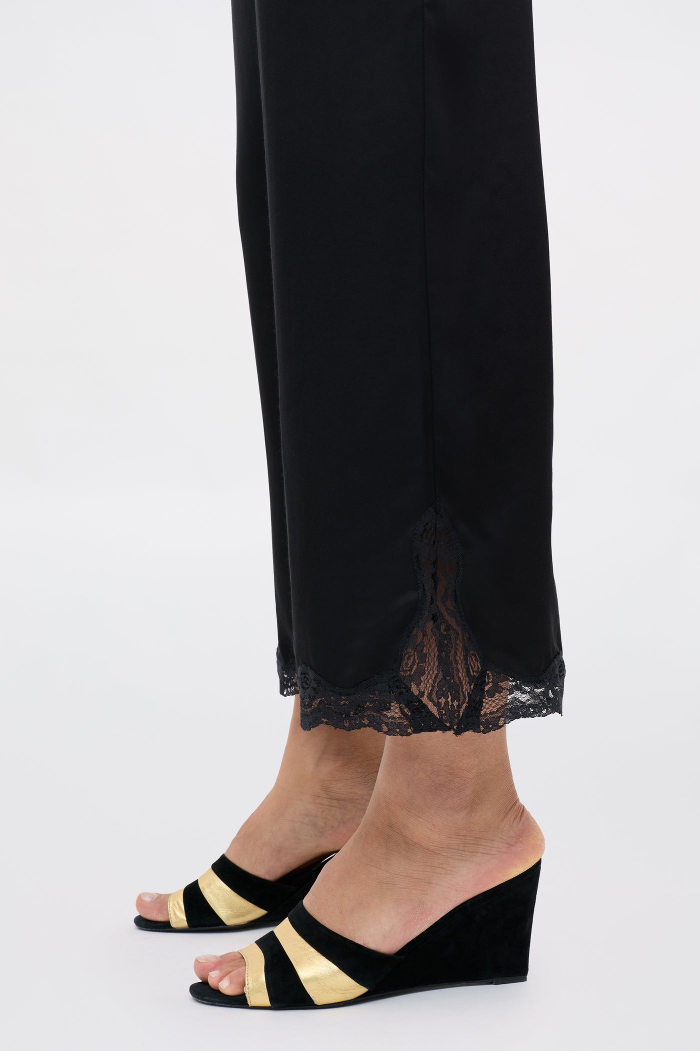 A person wearing the Mireille - Black satin pants with lace-trimmed hems and black wedge sandals featuring gold stripes, standing on a light background.