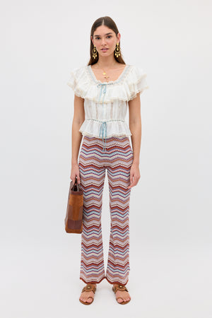 Wearing the Boni - Maria Chevron Multi patterned flared pants, a woman stands against a plain white background. She pairs these statement pants with a ruffled white cotton blouse, brown sandals, eye-catching earrings, and carries a brown handbag. This look is perfect for building a versatile wardrobe full of style and comfort.