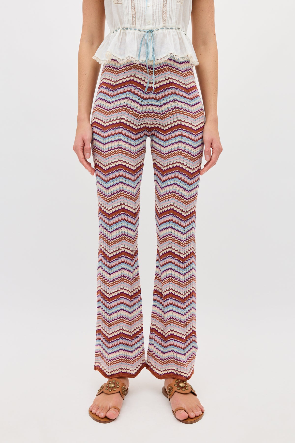 A person wearing the Boni - Maria Chevron Multi knitted trousers featuring a red, white, and purple zigzag pattern, styled with a white top with blue tie detail made from breathable cotton and brown sandals, standing against a plain white background.