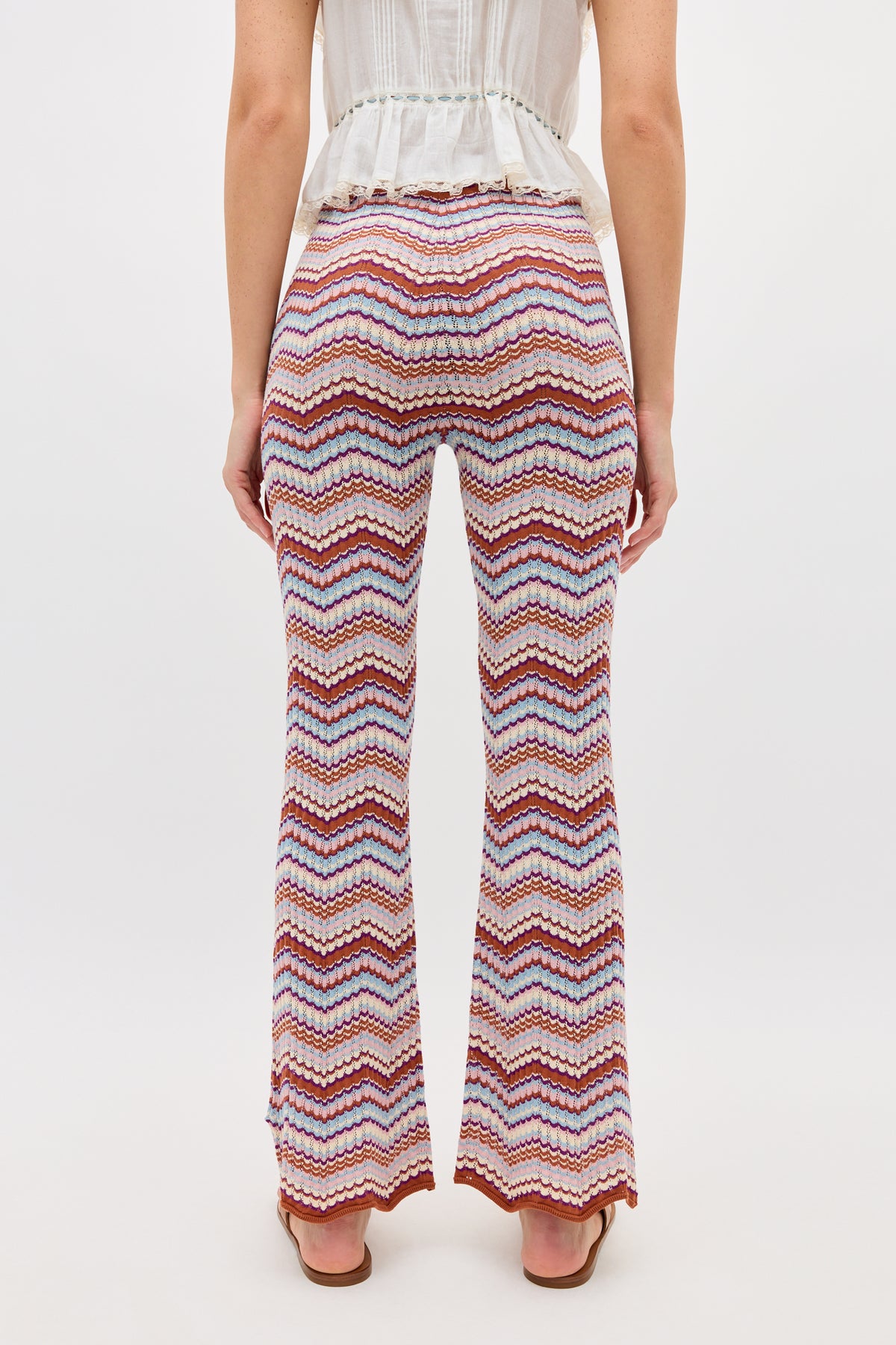 A person stands facing away, wearing a white sleeveless top and the Boni - Maria Chevron Multi trousers featuring a colorful zigzag knit pattern and flared legs. The background is plain white.
