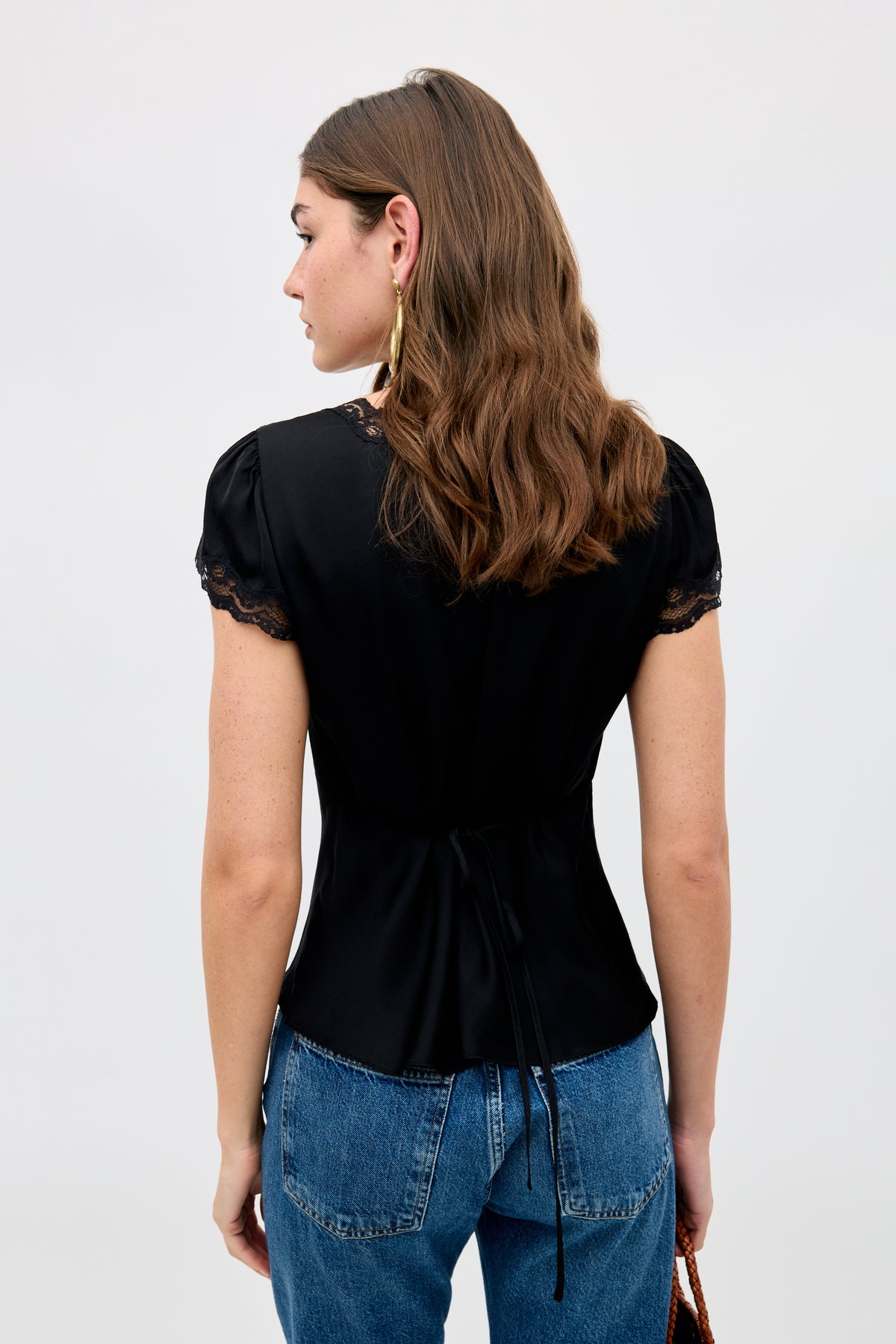 A woman with wavy brown hair stands facing away, wearing the Karia - Black short-sleeve blouse with lace trim and blue jeans, against a plain light background.