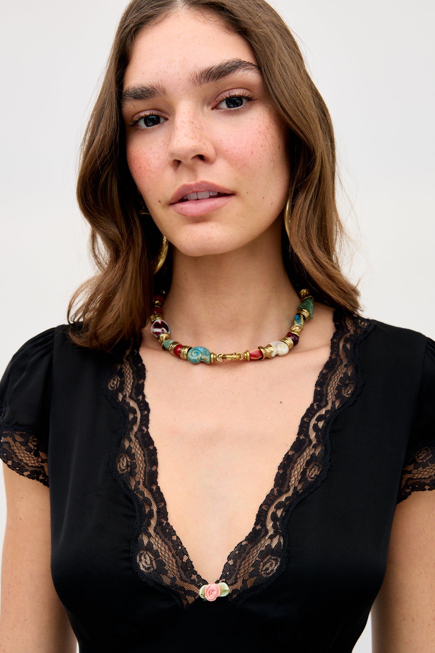 Woman with wavy brown hair wearing the Karia - Black lace-trimmed top and a colorful bead necklace, looking at the camera against a plain background.