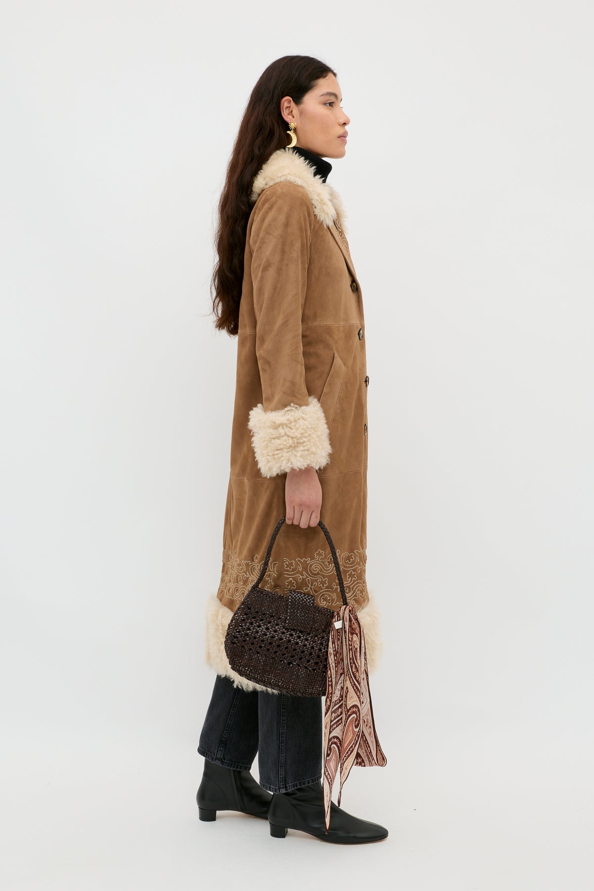 A woman in profile stands against a plain background, wearing the Cienna - Camel long tan shearling coat with white fur trim, dark jeans, black heeled shoes, and holding a woven dark handbag with a patterned scarf.