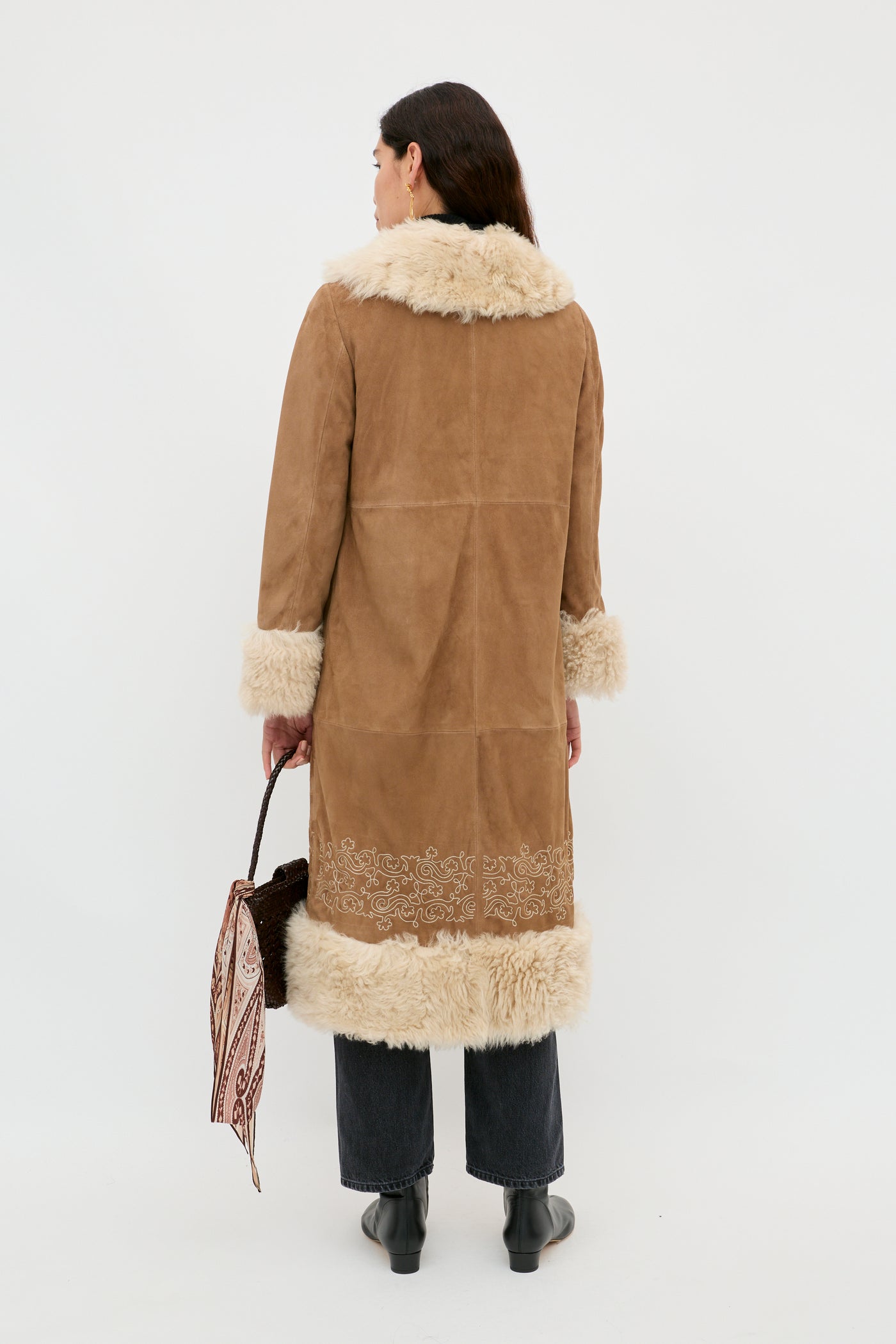 A woman with dark hair, seen from behind, wears the Cienna - Camel shearling coat with fur trim on the collar, cuffs, and hem. She pairs it with dark jeans, black boots, and carries a patterned handbag for a timeless look.