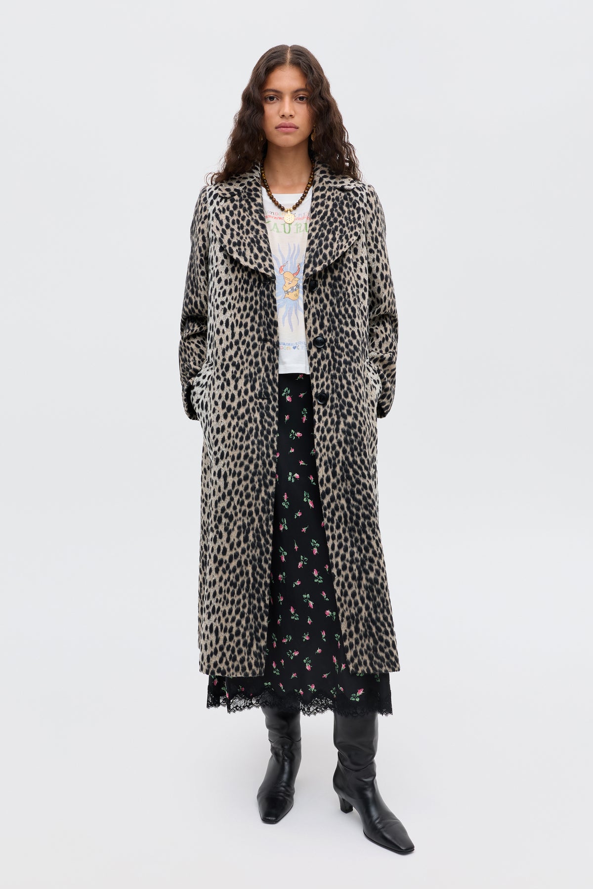 A woman with long, wavy hair and a neutral expression stands against a plain background wearing the Rosemary - Antelope Spot Jacquard Black coat over a white graphic tee, black floral skirt, and black knee-high boots.