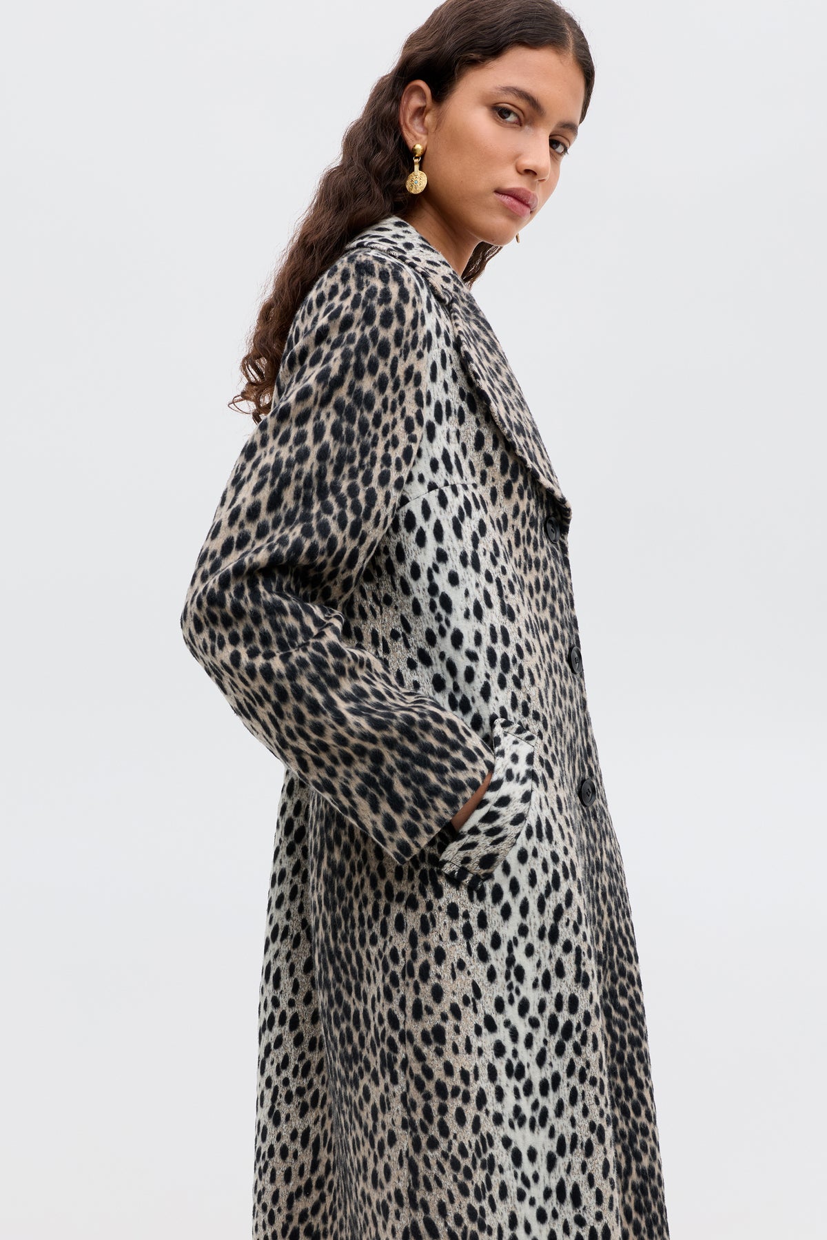 A woman with long curly hair wears the Rosemary - Antelope Spot Jacquard Black coat with curved lapels and gold earrings, posing with hands in her pockets against a plain light background.
