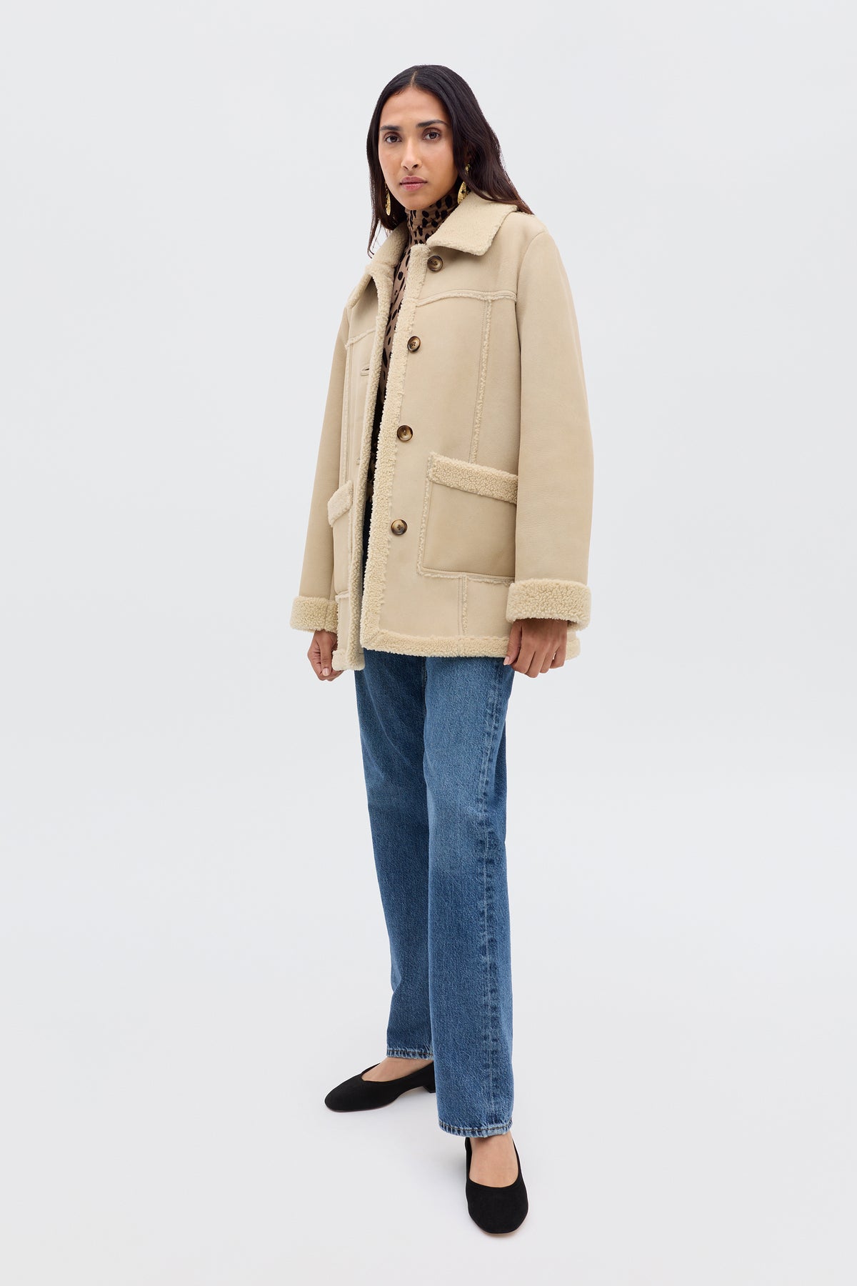 A person stands facing forward, wearing the Ellis - Cream beige faux-shearling jacket over a patterned top, blue jeans, and black shoes against a plain white background.