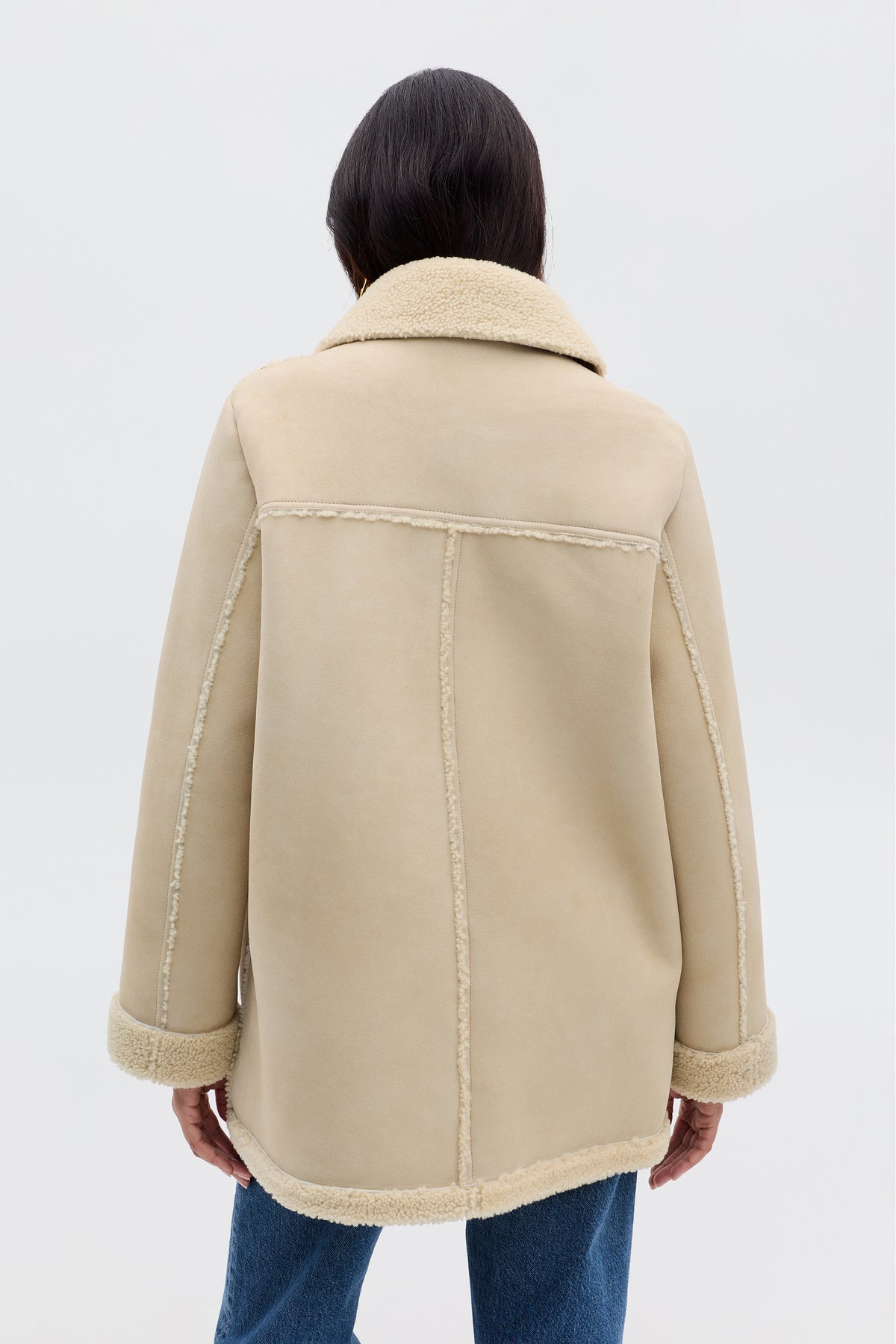 A person faces away, wearing Ellis - Cream: a beige faux-shearling coat with visible seams and a wide collar, paired with blue jeans against a plain, light background.